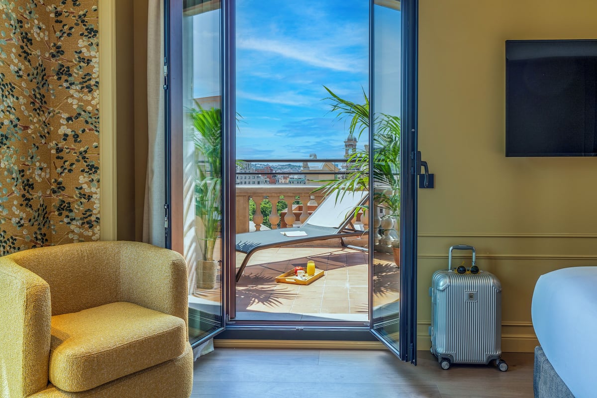 Une chambre d’hôtel dotée d’une grande porte-fenêtre coulissante donnant sur un balcon avec vue sur la ville, un lit, une chaise et une valise.