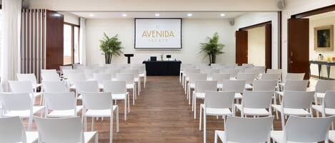 A conference room with rows of white chairs, a podium, and a projector screen displaying "EL AVENIDA PALACE".