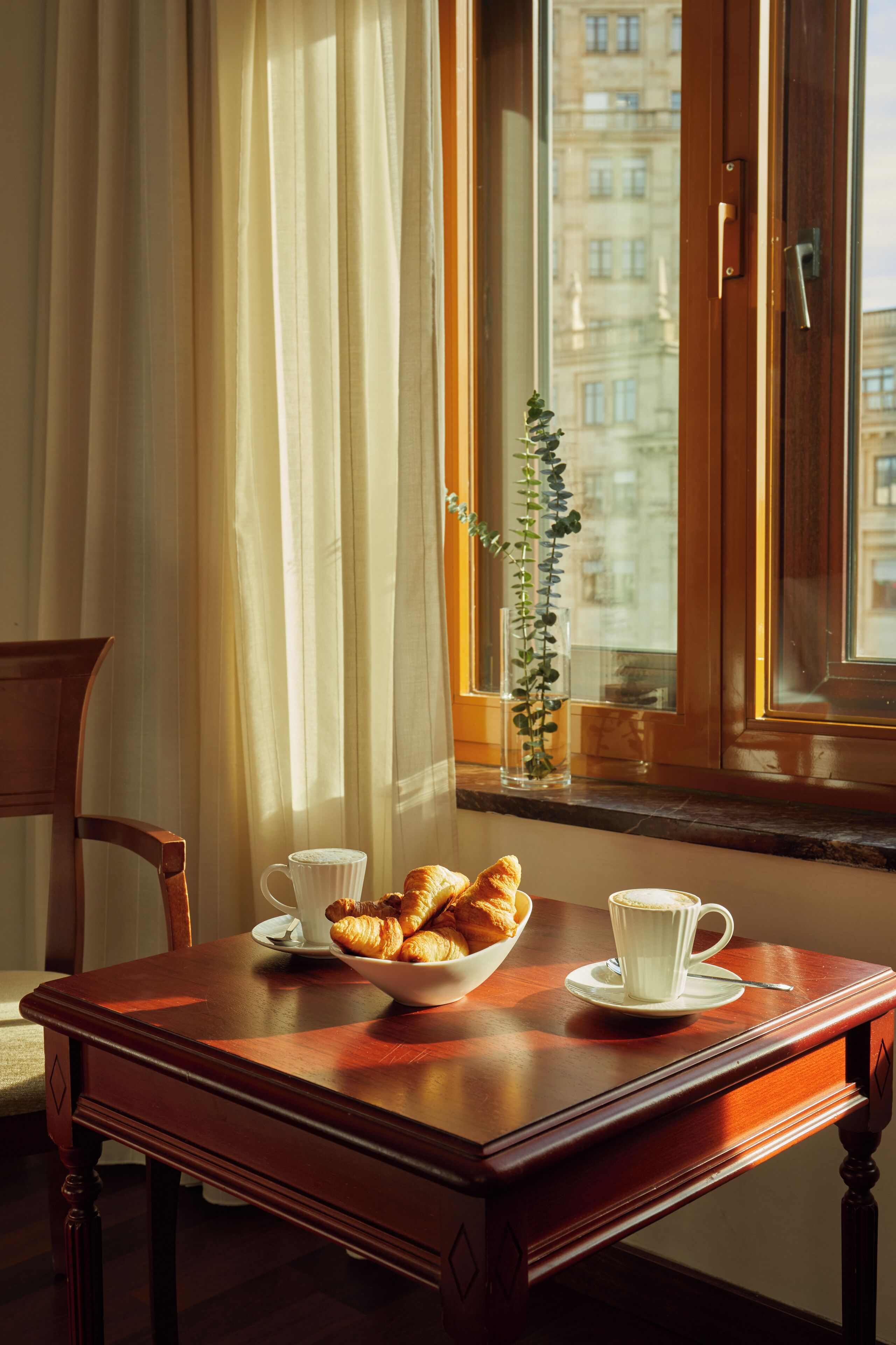 A wooden table with a bowl of croissants and two white cups, a potted plant, and a window with curtains.