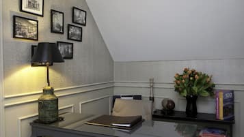 A corner office with a desk, lamp, framed pictures, a vase with flowers, and books.