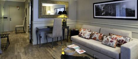 A modern living room with a grey sofa, a glass coffee table, a desk with a chair, a large mirror, and a framed picture on the wall.
