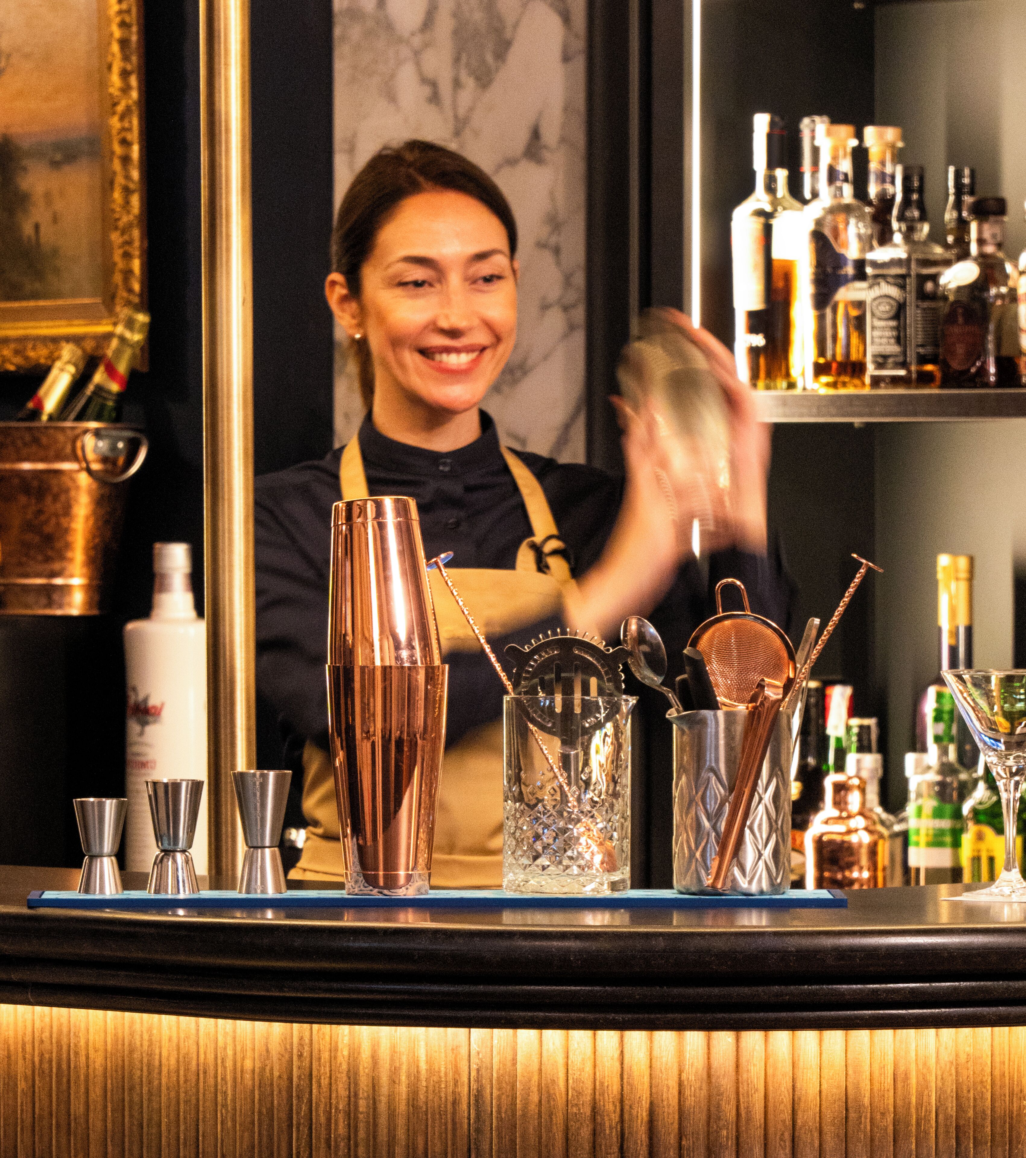 Uma área de bar com um barman, diversas ferramentas de bar e garrafas de bebidas alcoólicas.