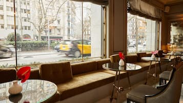 Un rincón acogedor con sofá y mesas, amplias ventanas con vista a la calle, y una lámpara sobre la mesa.
