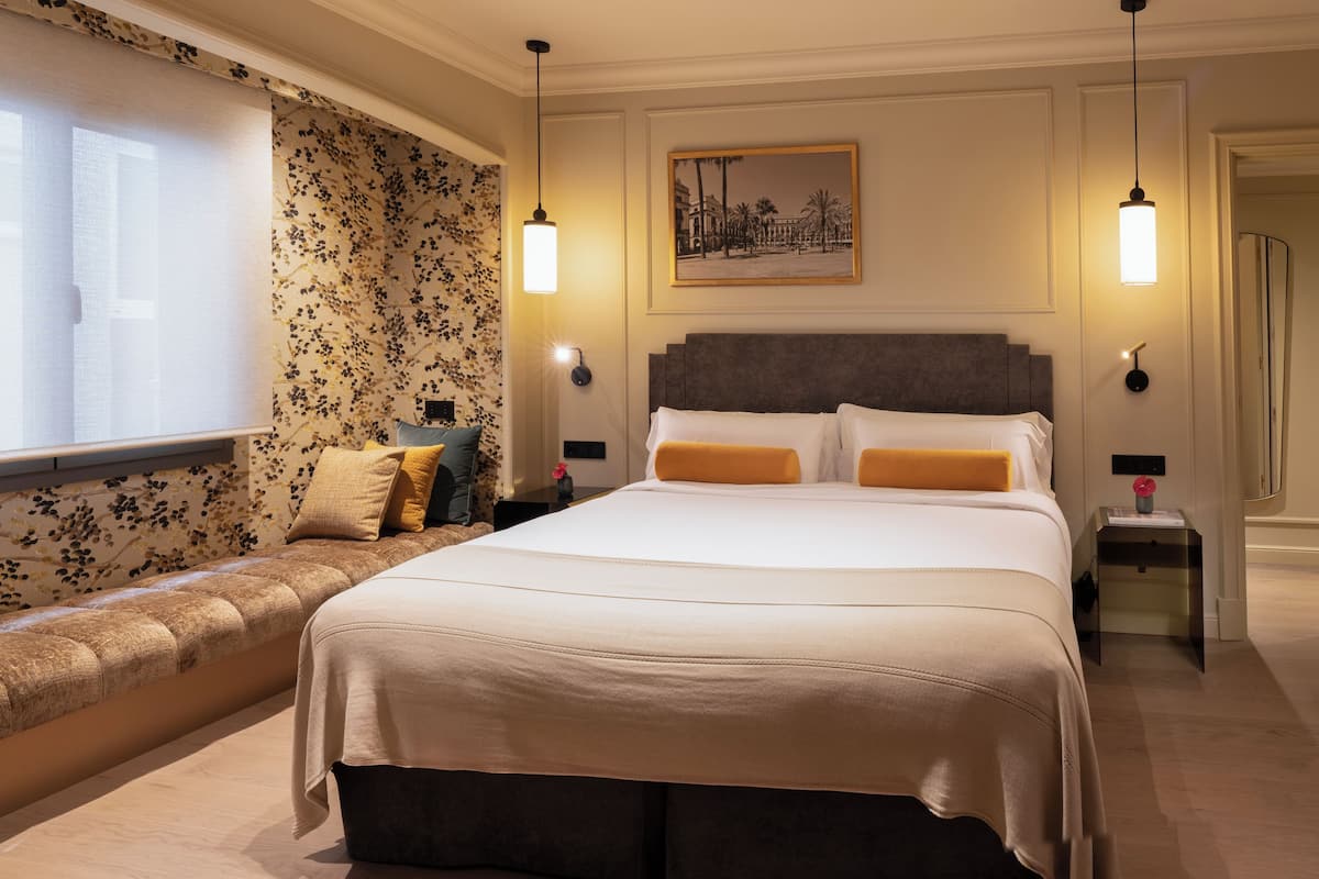 Une chambre d’hôtel avec un grand lit, une banquette avec des coussins, un tableau accroché au mur et deux lampes de chevet.