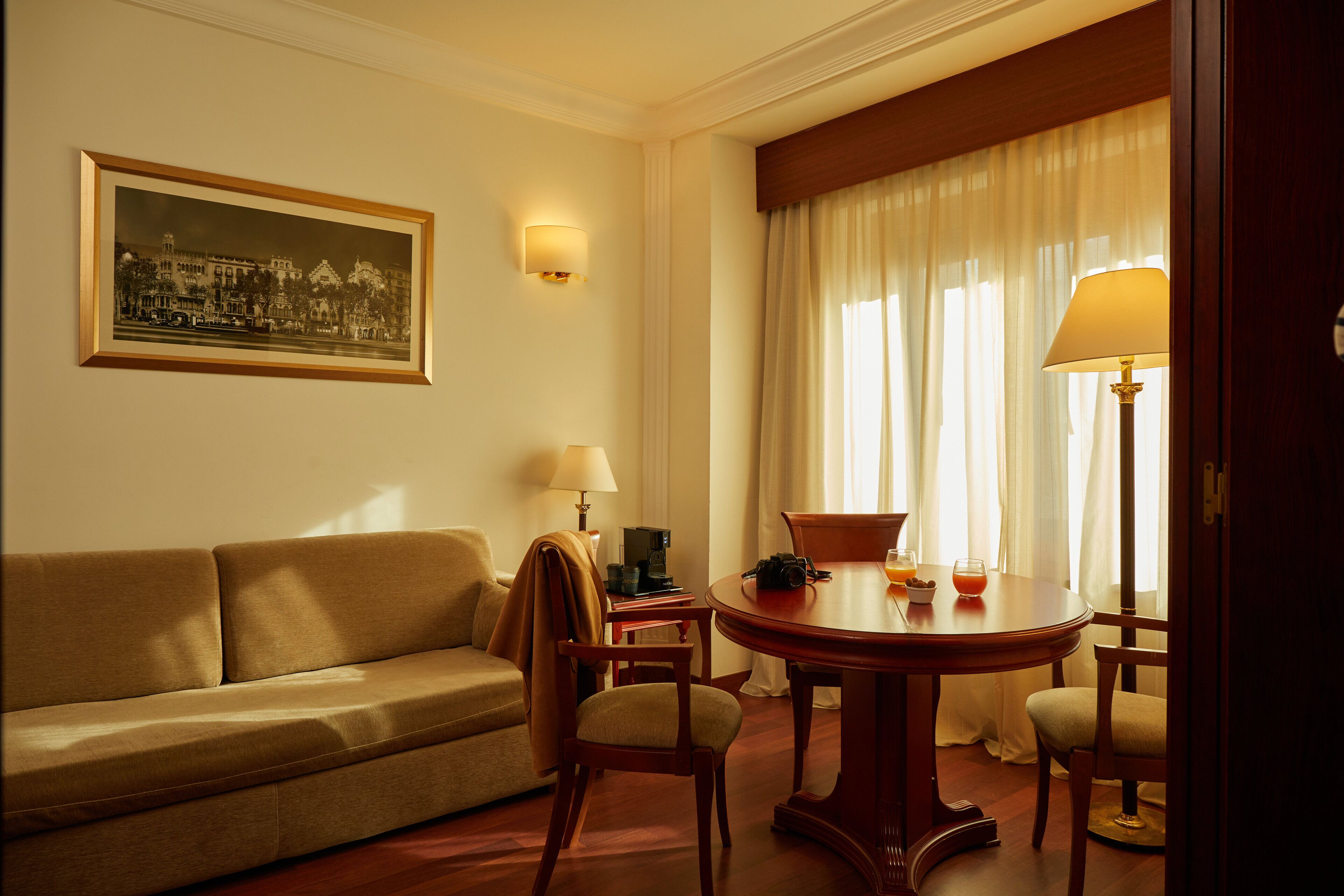 Habitación de hotel con sofá, una mesa de comedor con sillas, lámpara, un cuadro en la pared y una ventana con cortinas.