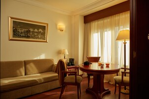 A hotel room with a sofa, a dining table with chairs, a lamp, a painting on the wall, and a window with curtains.