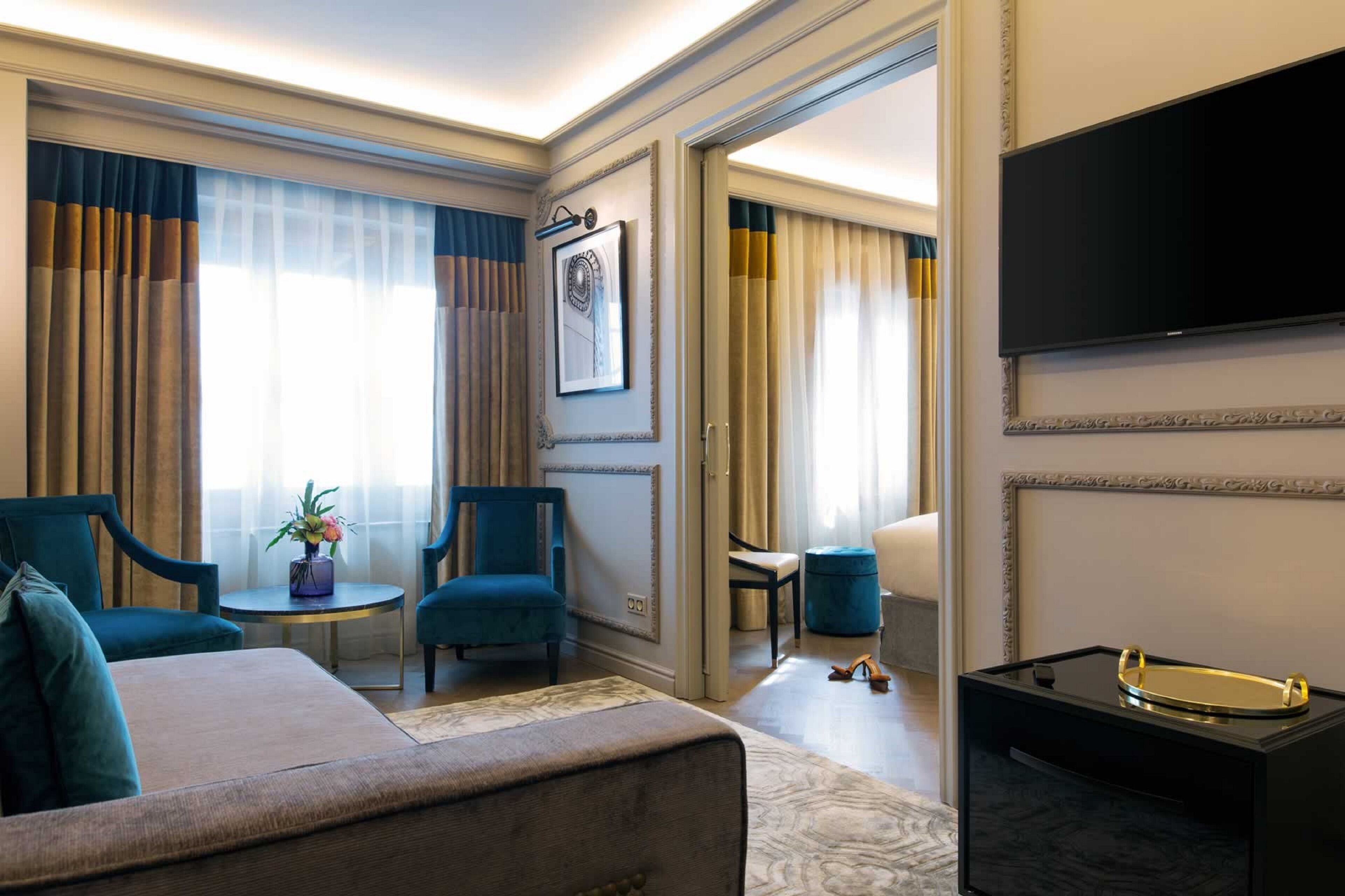 A hotel room with a bed, two armchairs, a small table with a plant, a TV, and a minibar.