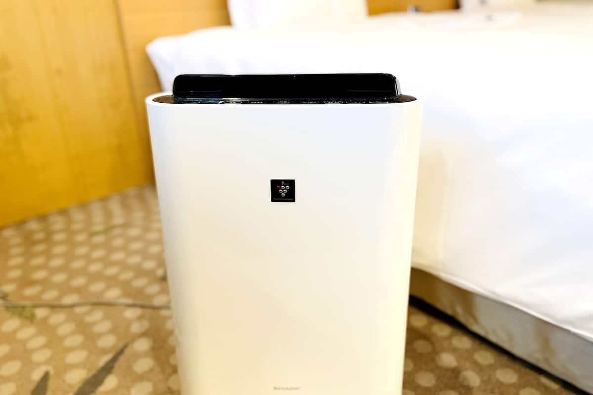 A white, modern air purifier with a small logo on the front, placed on a patterned carpet in a hotel room.