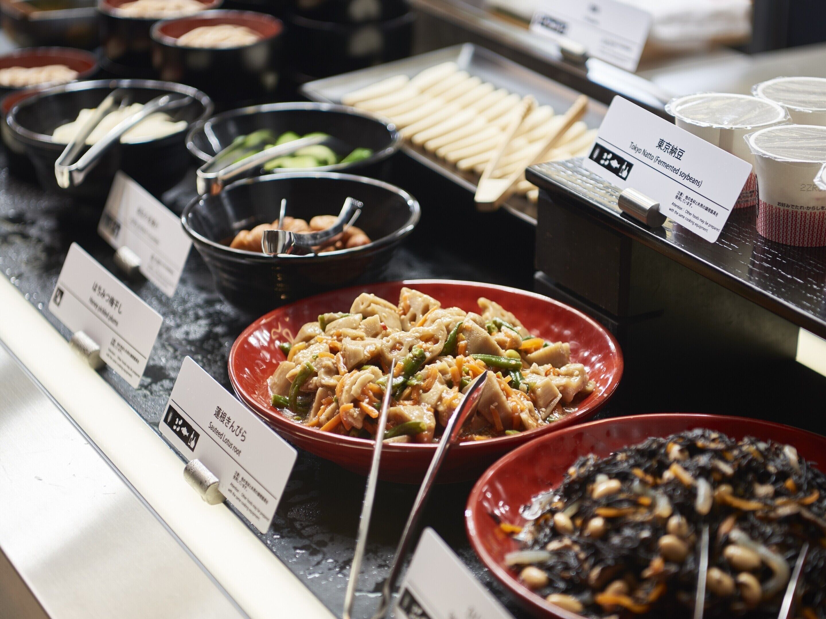 A buffet with various dishes including a red bowl of stir-fried vegetables, a black bowl of tofu, and a tray of breadsticks.