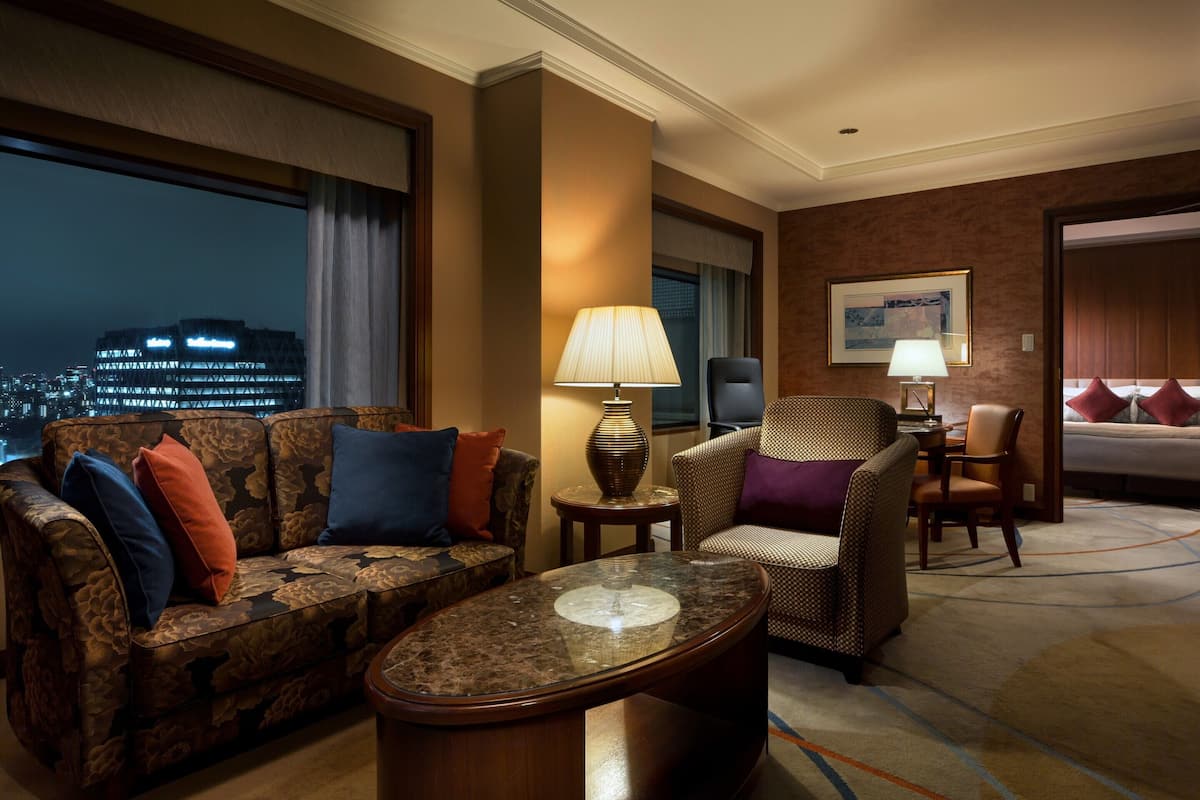 A hotel room with a sofa, armchair, and a small table. There is a window with a view of a cityscape.