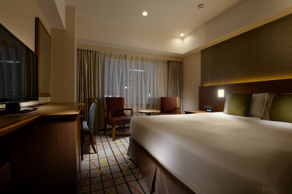 A hotel room with a large bed, a desk with a computer, a chair, a small table, and a window with curtains.