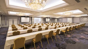 A conference room with rows of tables and chairs, large chandeliers, and a carpeted floor.