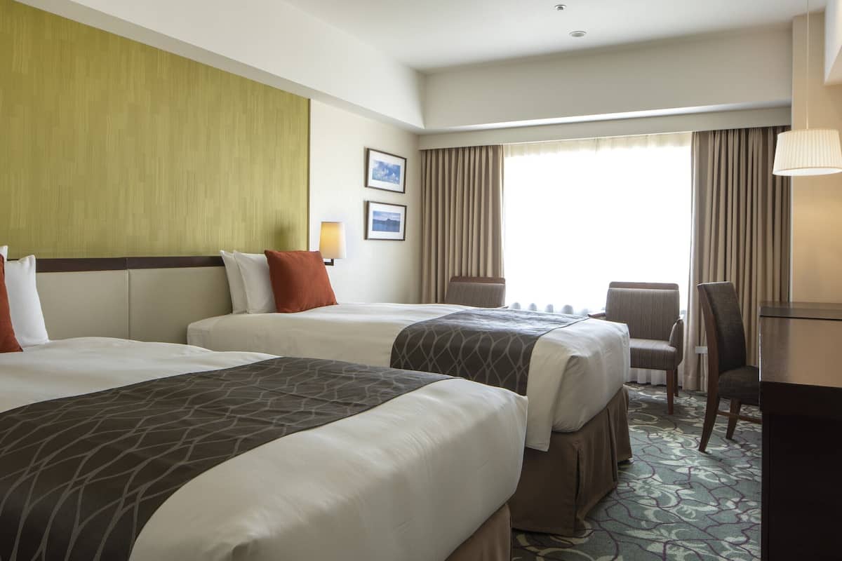 A hotel room with two beds, a desk, a chair, and a window with curtains.
