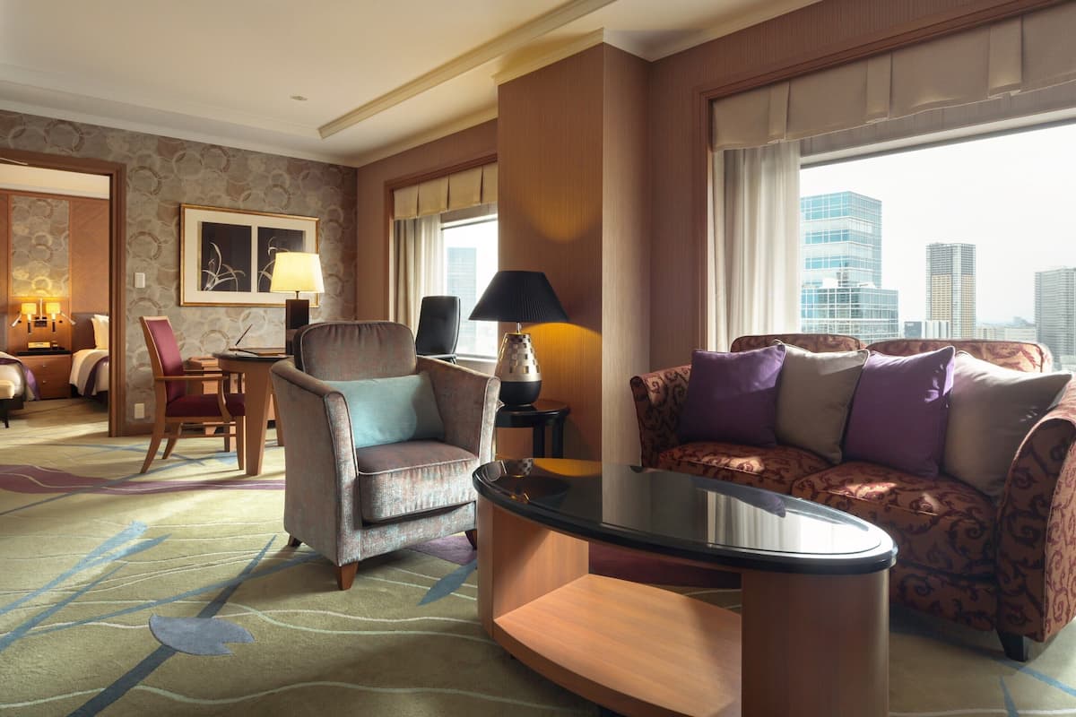 A hotel room with a city view, a sitting area with a sofa and armchairs, a dining table, and a bed with a headboard.