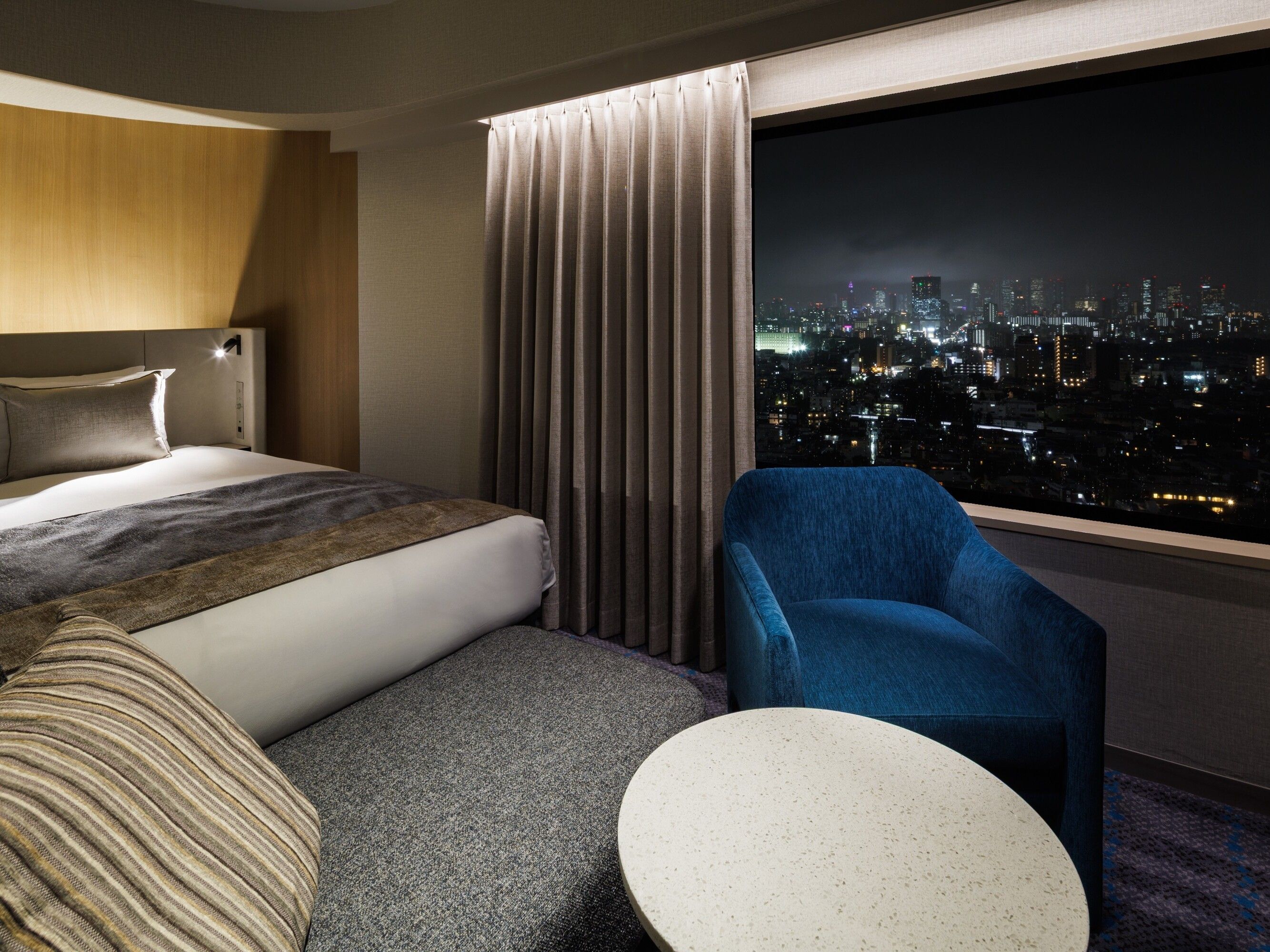 A hotel room with a bed, a blue armchair, a small round table, and a view of a cityscape at night.