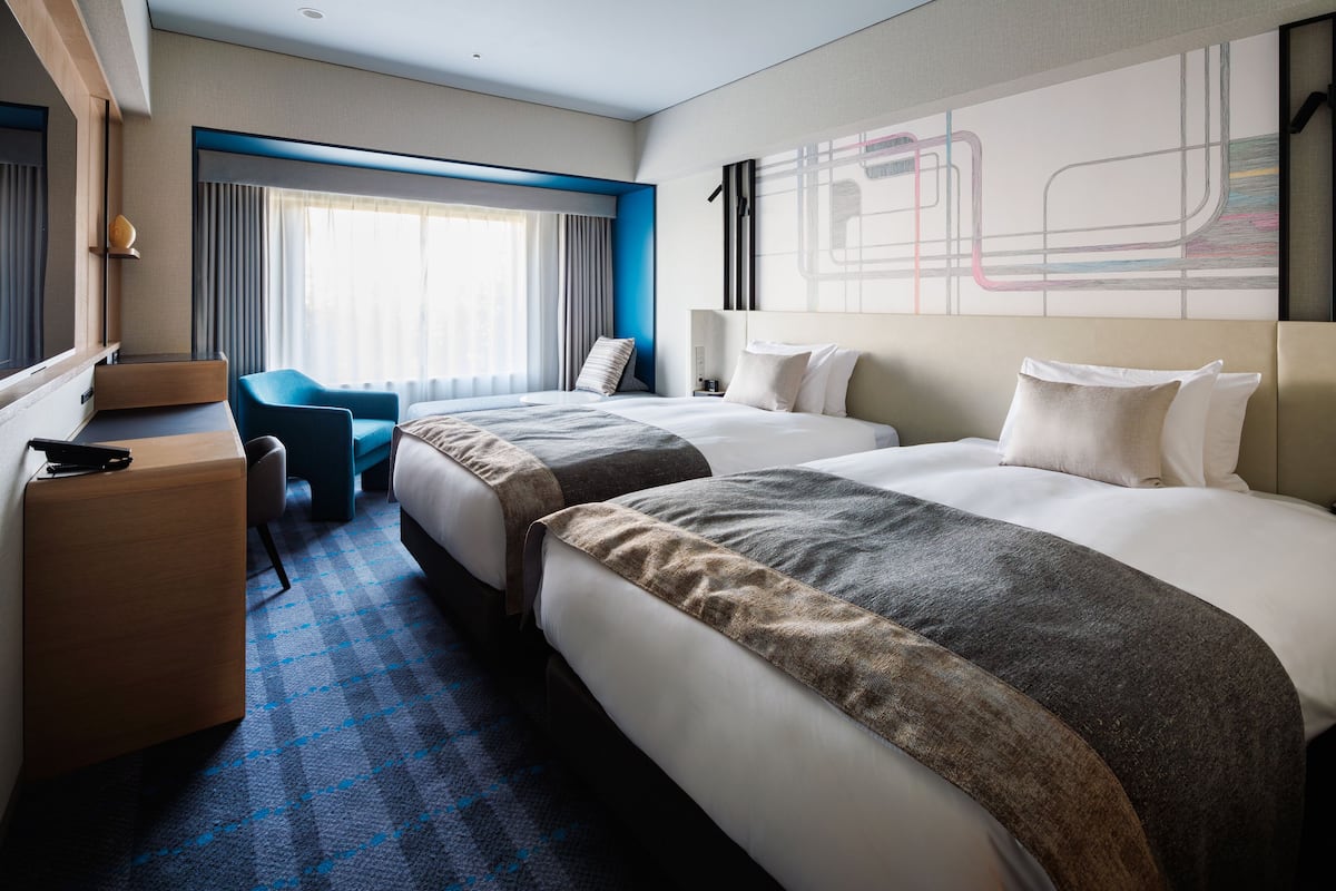 A hotel room with two beds, a desk, a chair, and a large window with curtains.