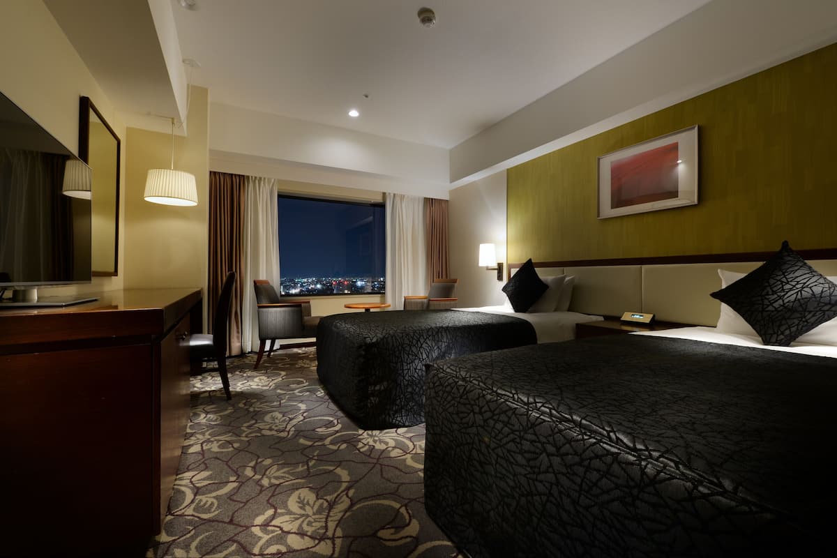 A hotel room with two beds, a desk, a chair, a lamp, and a view of the city at night.