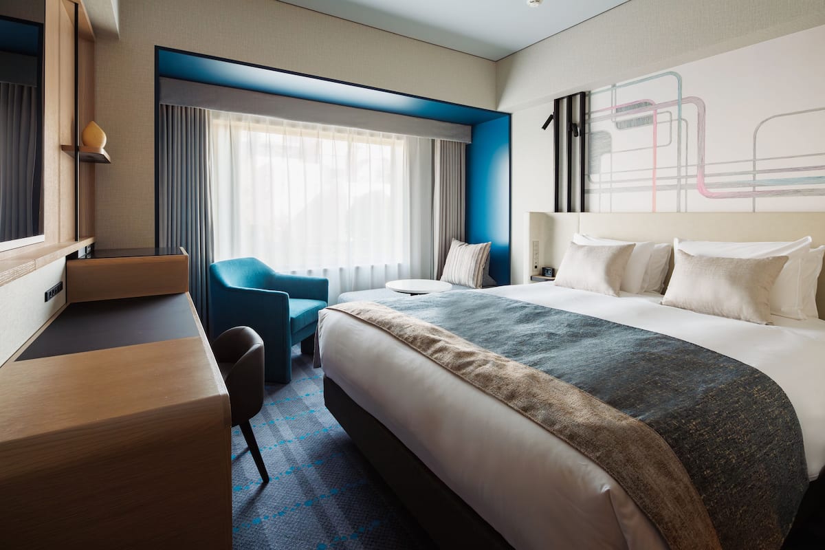 A modern hotel room with a large bed, a desk, a chair, and a window with curtains.