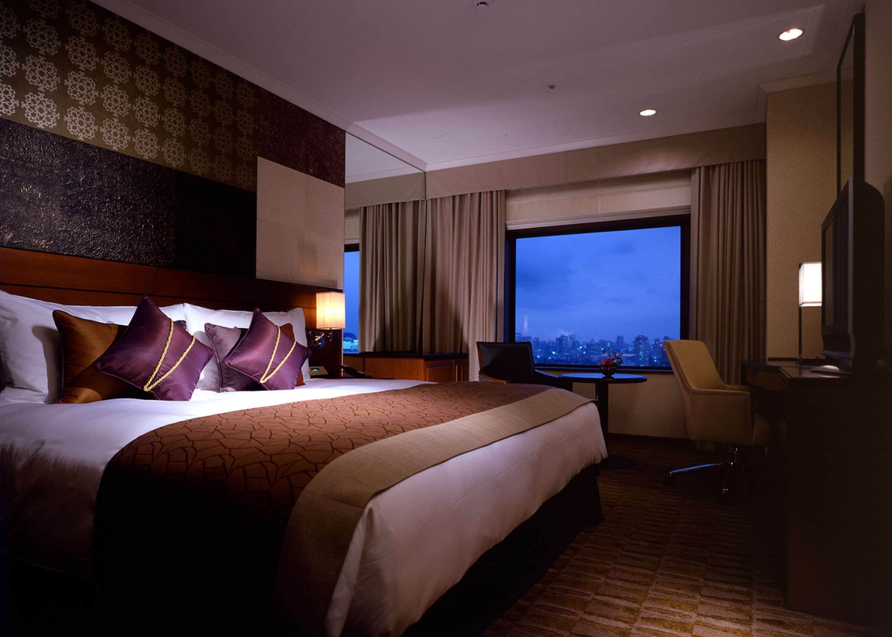 A hotel room with a large bed, a desk, a chair, a TV, and a city view.