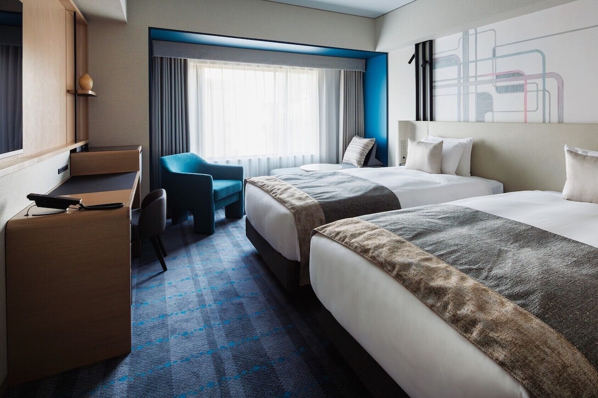 A hotel room with two beds, a desk, a chair, and a large window with curtains.