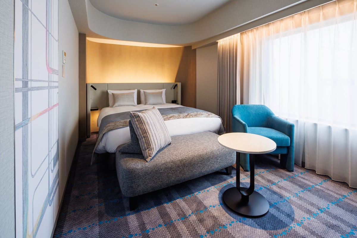 A modern hotel room with a large bed, a small sofa, a round table, and a large window with curtains.