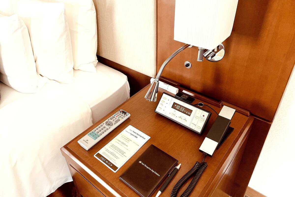 A hotel room with a bed, a bedside table with a digital clock, a remote control, a pen, and a notebook.