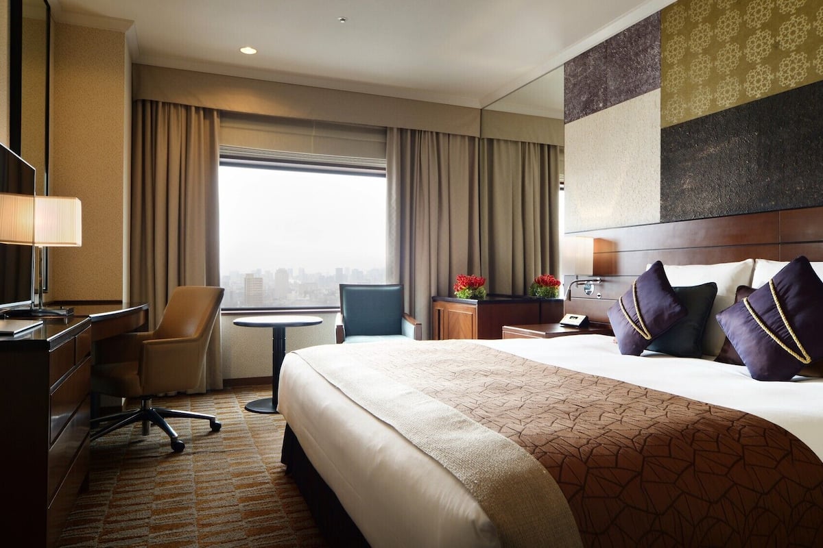 A hotel room with a bed, desk, chair, TV, and a city view.