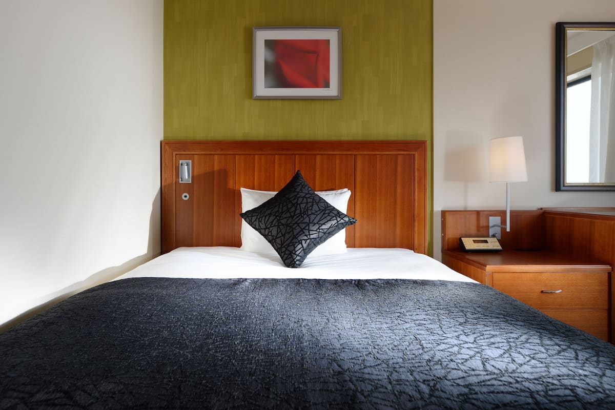 A hotel room with a wooden headboard, a bed with a black textured bedspread and a geometric pillow, a bedside table with a lamp, and a framed picture on the wall.