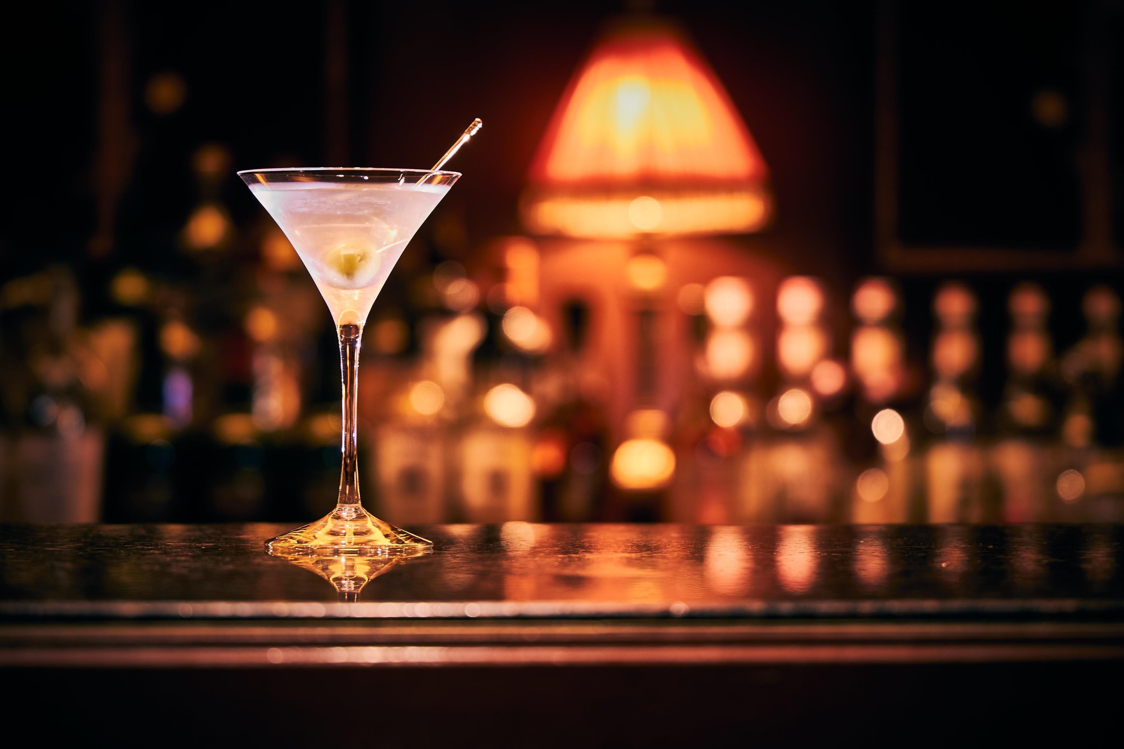 Una copa de martini sobre una barra, con un fondo desenfocado de botellas y una luz cálida.