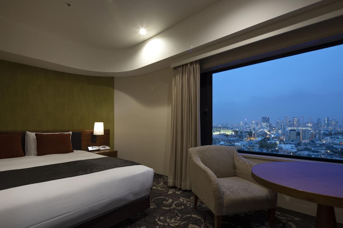 A hotel room with a bed, a chair, a table, a nightstand, and a view of a cityscape at dusk.