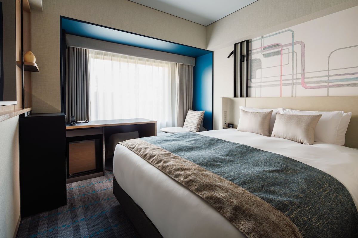 A hotel room with a bed, a desk, a chair, and a large window with curtains.