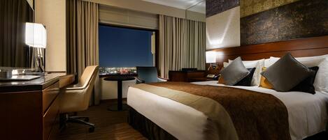A hotel room with a bed, desk, chair, and a view of the city at night.