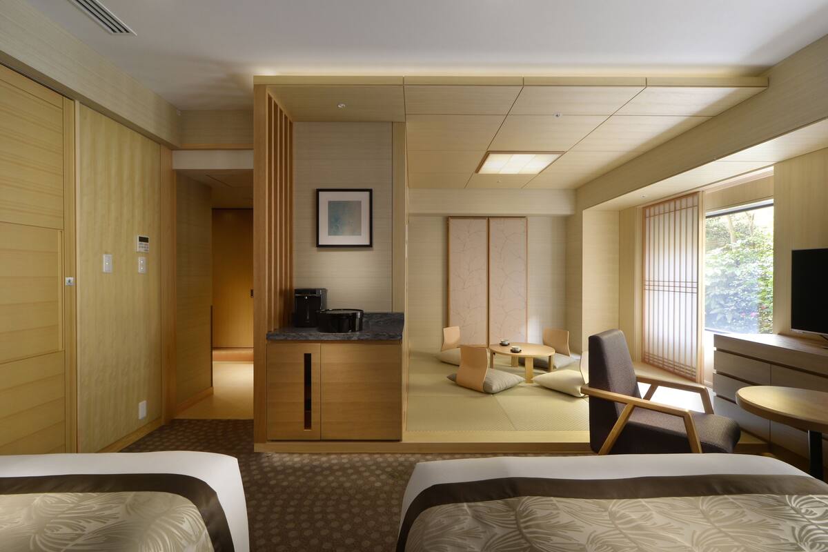 A hotel room with two beds, a small kitchenette, a sitting area with a tatami mat, and a large window offering a view of greenery.