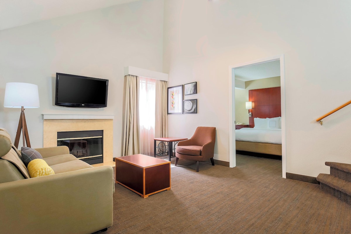 A hotel room with a sofa, armchair, small table, flat-screen TV, fireplace, and a staircase leading to another room.