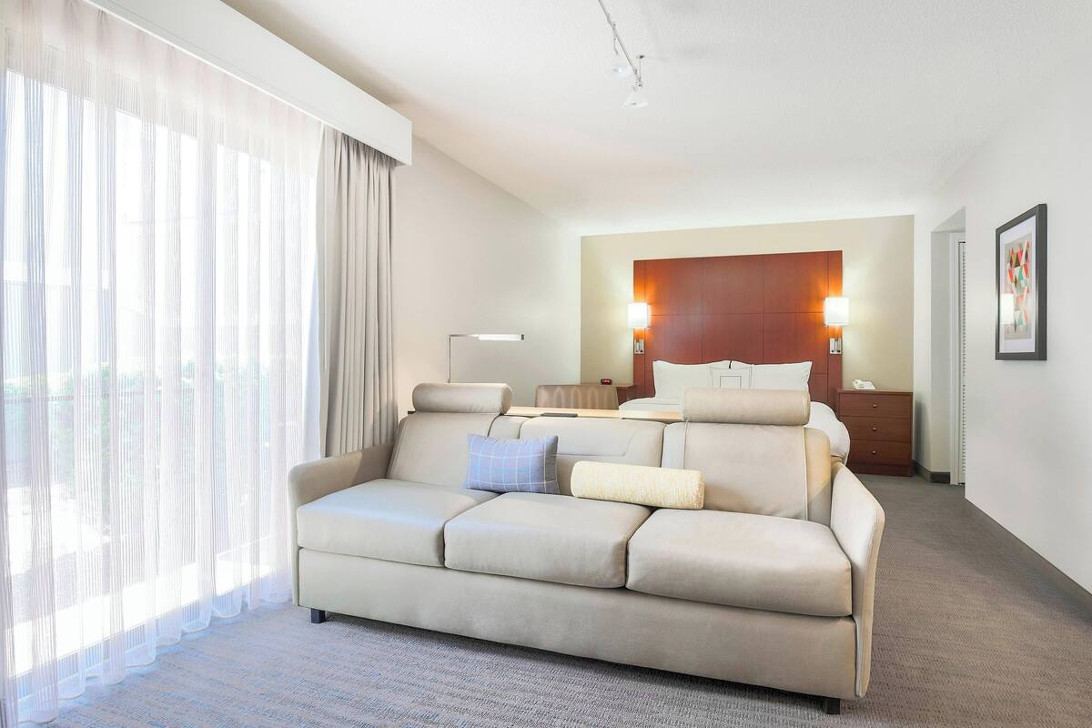 A hotel room with a sofa, a bed, a nightstand, and a large window with sheer curtains.