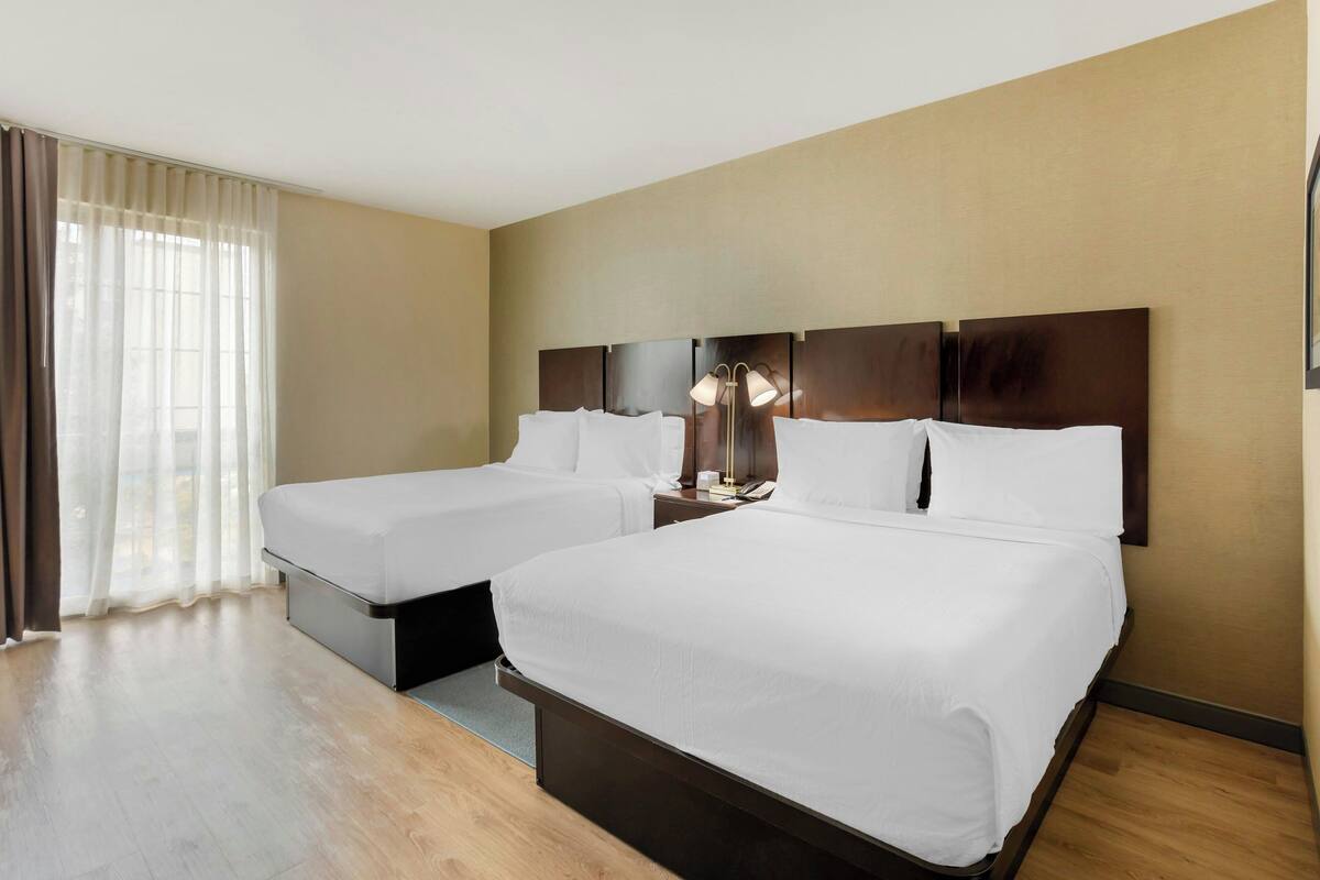 A hotel room with two beds, a nightstand with a lamp, and a large window with sheer curtains.