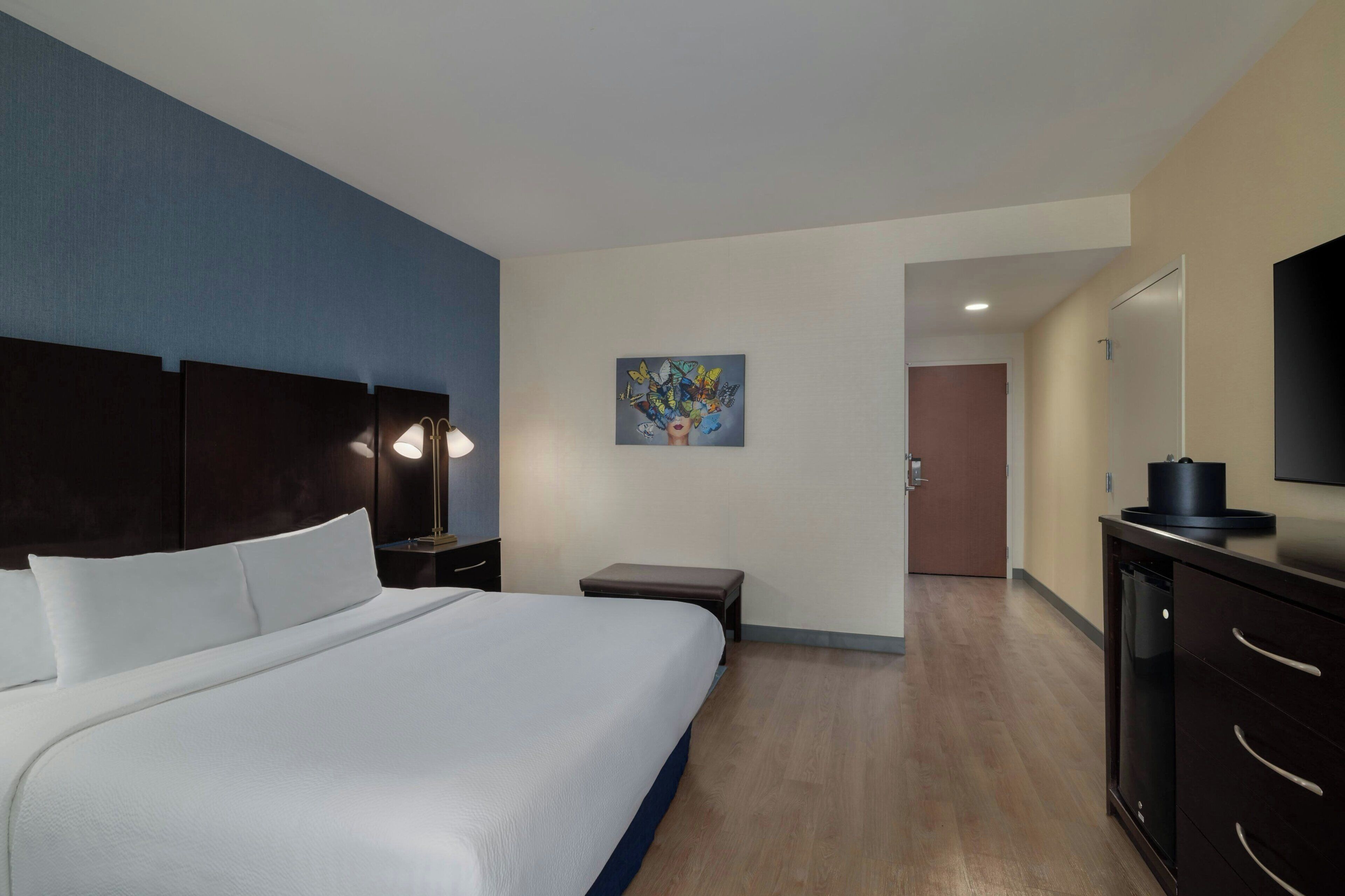 Quarto de hotel com uma cama grande, uma mesinha de cabeceira com abajur, um pequeno banco, um quadro na parede, uma porta que dá para outro cômodo e uma televisão em um criado-mudo.