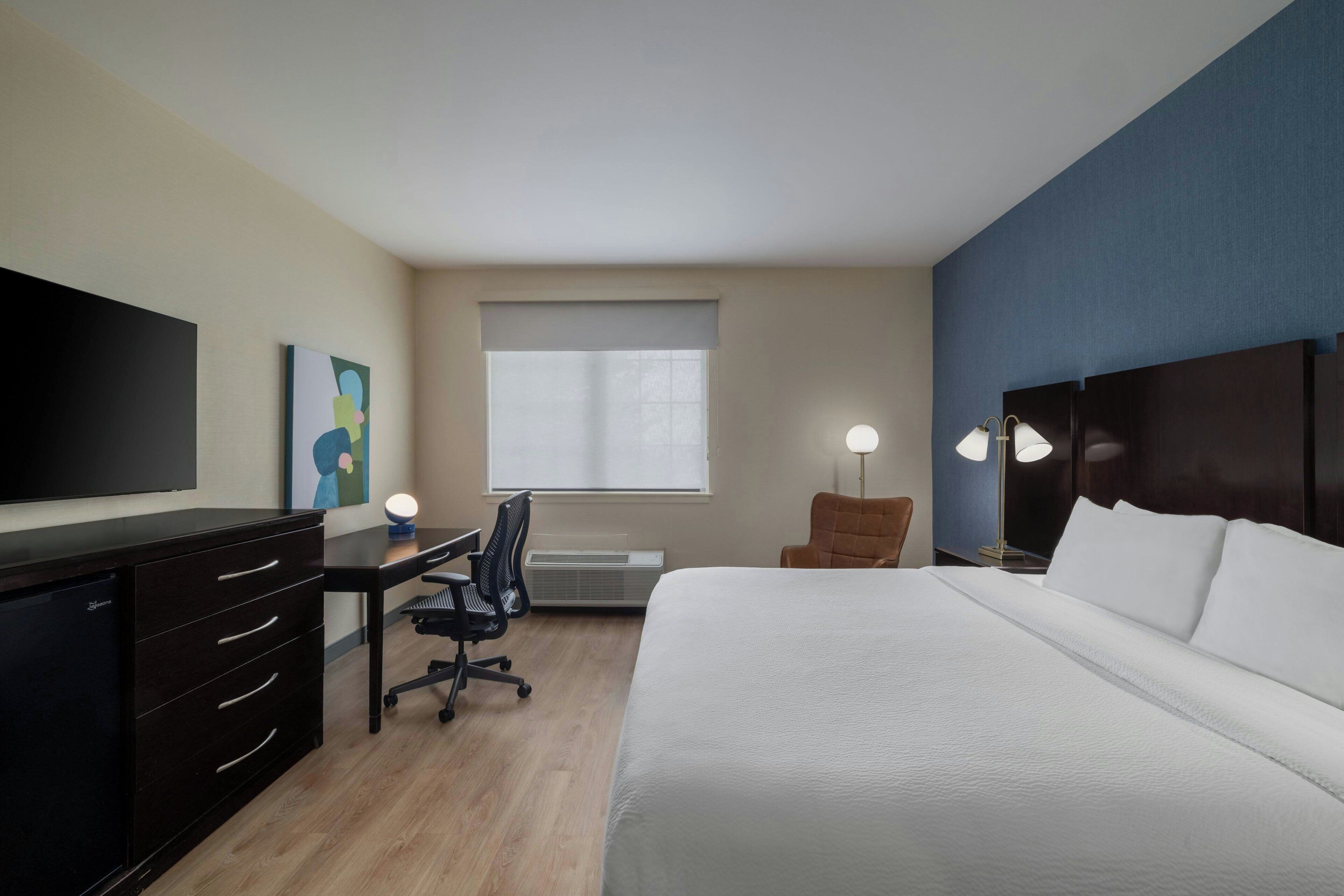 Una habitación de hotel con cama, escritorio, silla, televisión y una obra de arte en la pared.