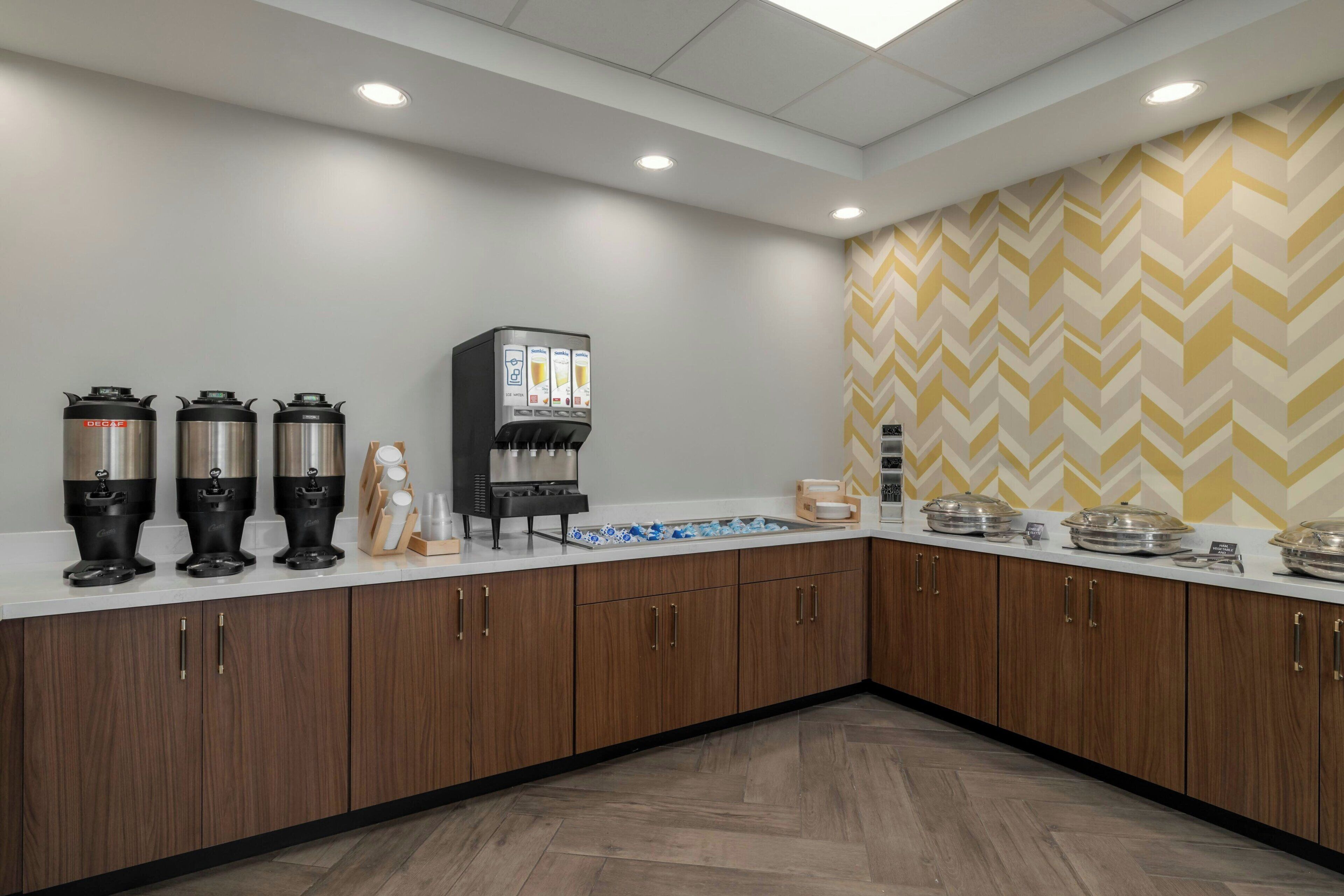 A modern kitchen area with a coffee station, a beverage dispenser, and a buffet setup.