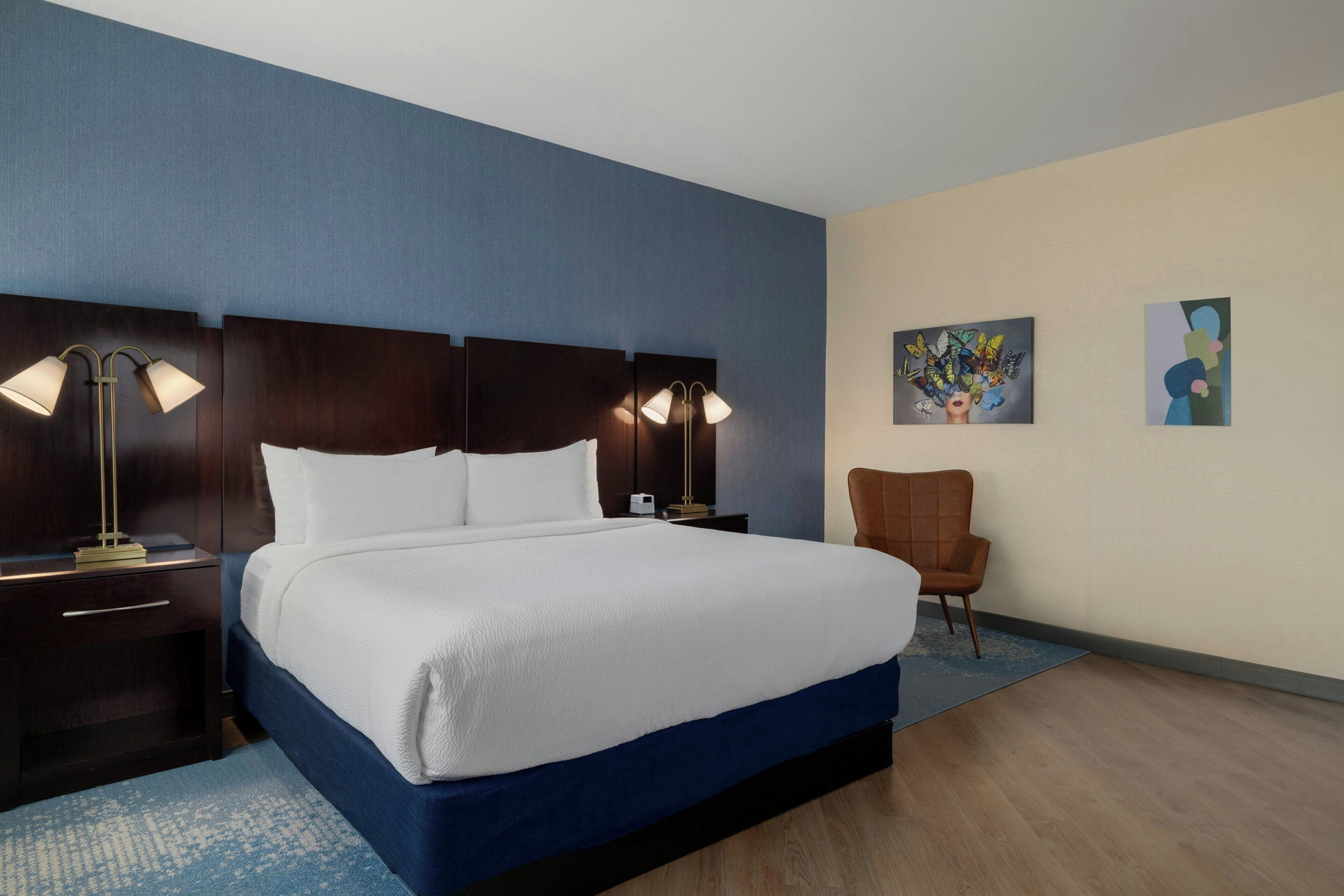 A hotel room with a large bed, two bedside tables with lamps, a chair, and two wall art pieces.