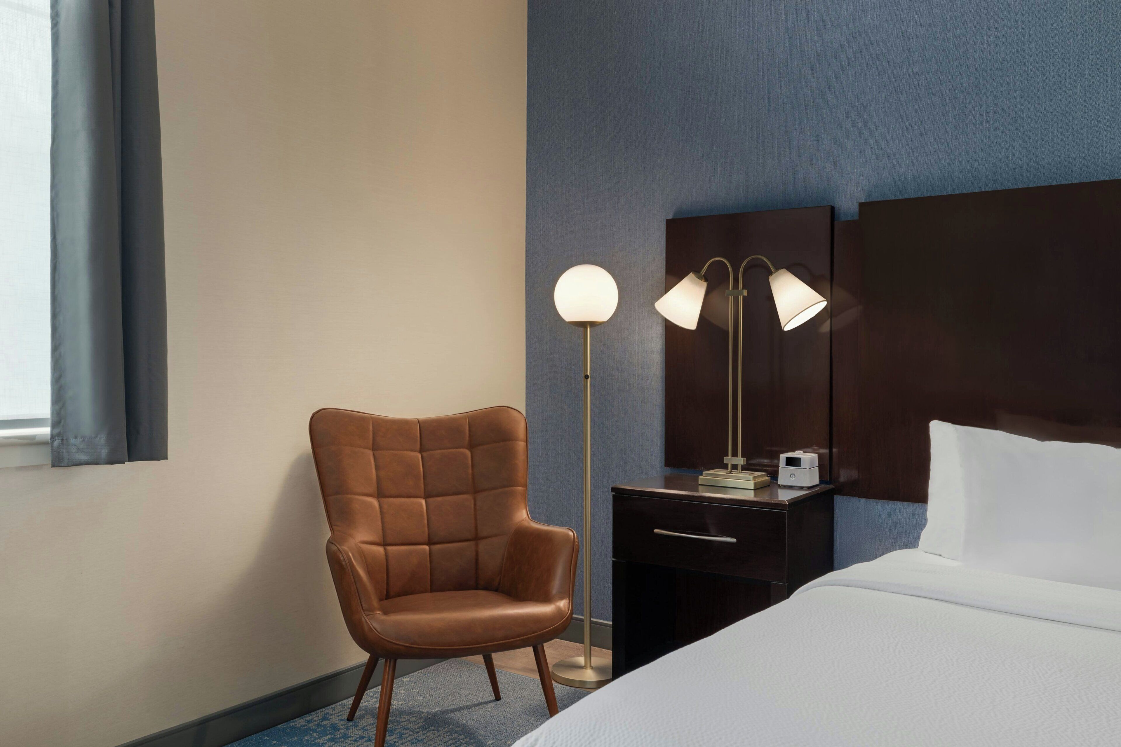 Ett hotellrum med en säng, en stol, ett skrivbord med en lampa och ett nattduksbord med en skrivare.