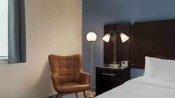 Habitación de hotel con cama, una silla, un escritorio con lámpara y una mesita de noche con una impresora.