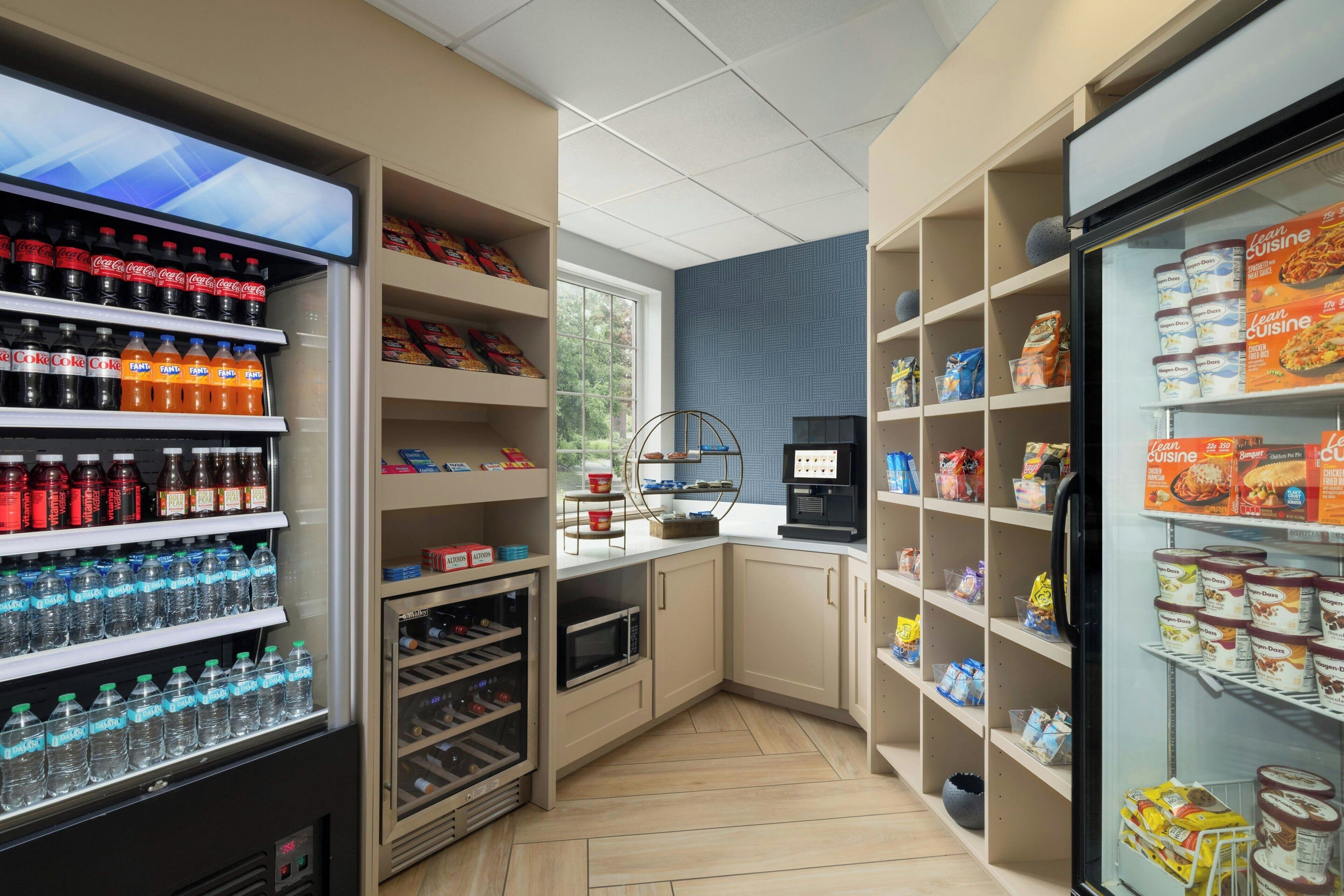 A kitchenette with a refrigerator stocked with food items, a microwave, a coffee machine, and shelves with bottled water and snacks.