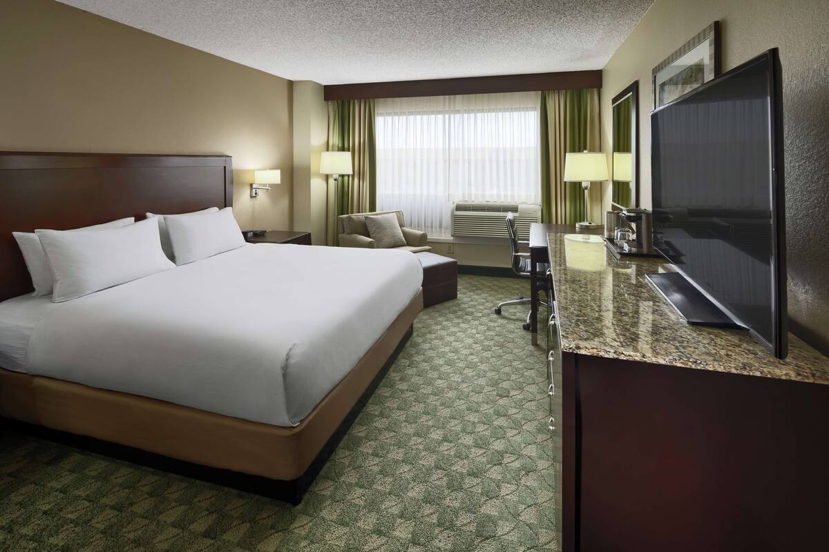 A hotel room with a large bed, a desk with a flat-screen TV, a chair, and a desk lamp.