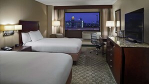 Habitación de hotel con dos camas, un escritorio, televisión y vistas a la ciudad iluminada por la noche.