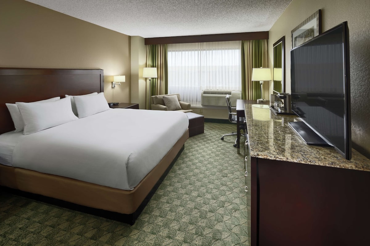 A hotel room with a large bed, a desk with a flat-screen TV, a chair, a desk lamp, and a window with curtains.