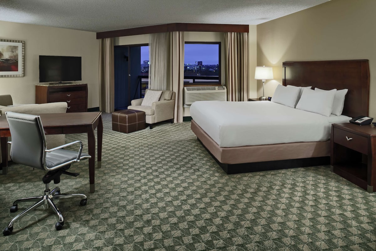 A hotel room with a large bed, a desk, a chair, a TV, and a view of the city at night.