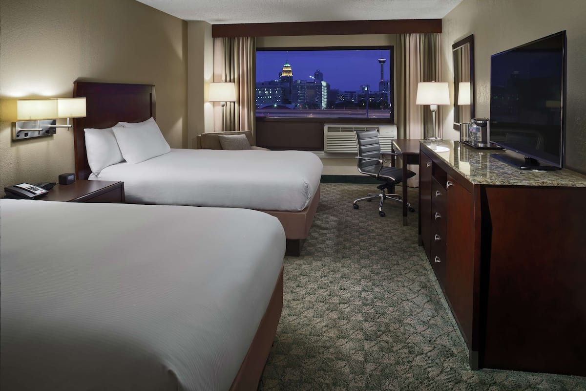 A hotel room with two beds, a desk, a television, and a view of a cityscape at dusk.