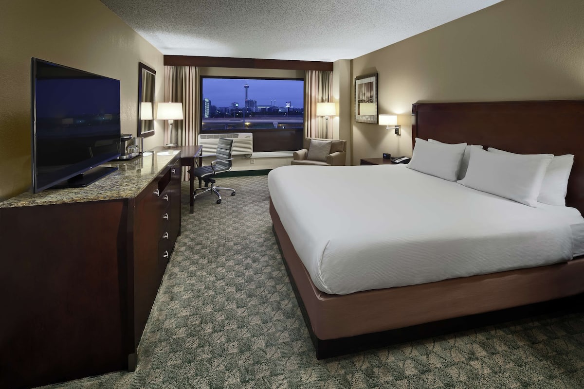 A hotel room with a large bed, a desk with a flat-screen TV, a chair, a desk lamp, and a view of a cityscape at dusk.