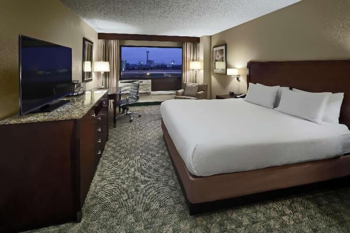 A hotel room with a large bed, a desk with a flat-screen TV, a chair, a desk lamp, and a view of a cityscape through a large window.
